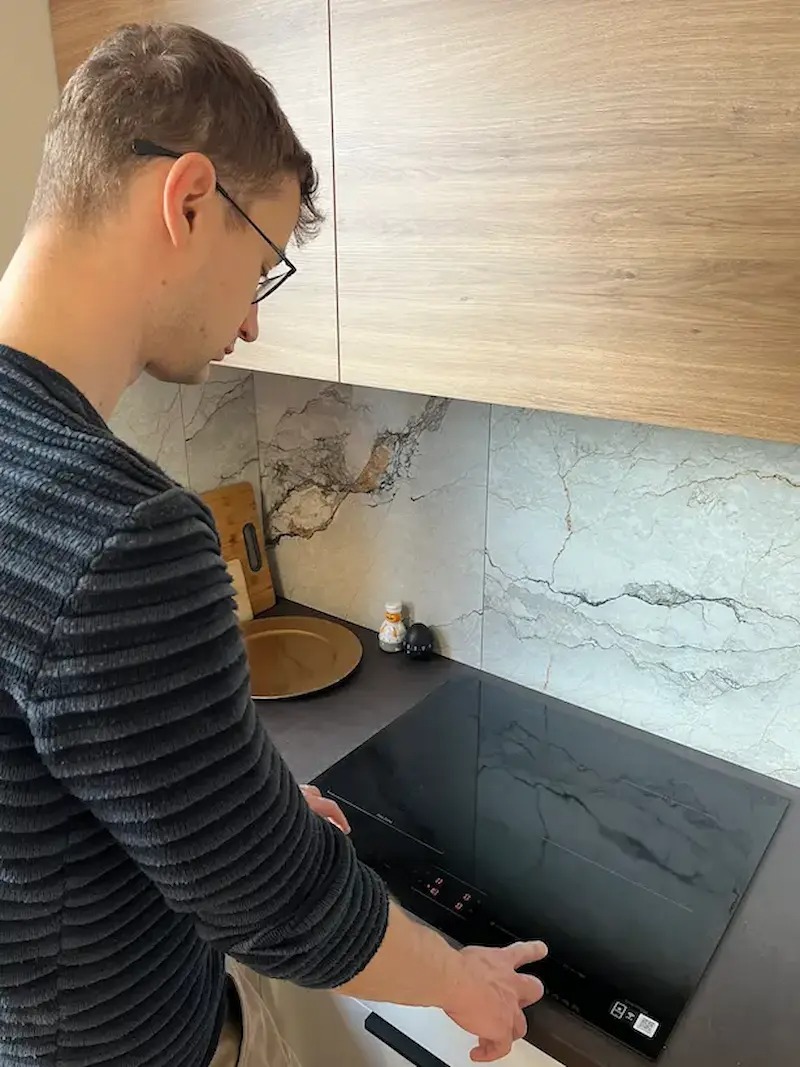 Mężczyzna w okularach włącza płytę indukcyjną w nowoczesnej kuchni z marmurowym backsplashiem i drewnianymi szafkami. Widoczne przyprawy i deska do krojenia.