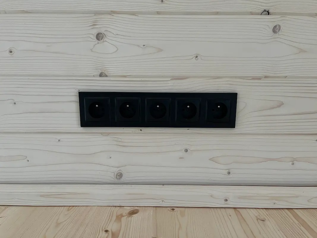 Rząd pięciu czarnych gniazdek elektrycznych zamontowanych na jasnej, drewnianej ścianie z widoczną strukturą desek. Widok z bliska, poziomy kadr.