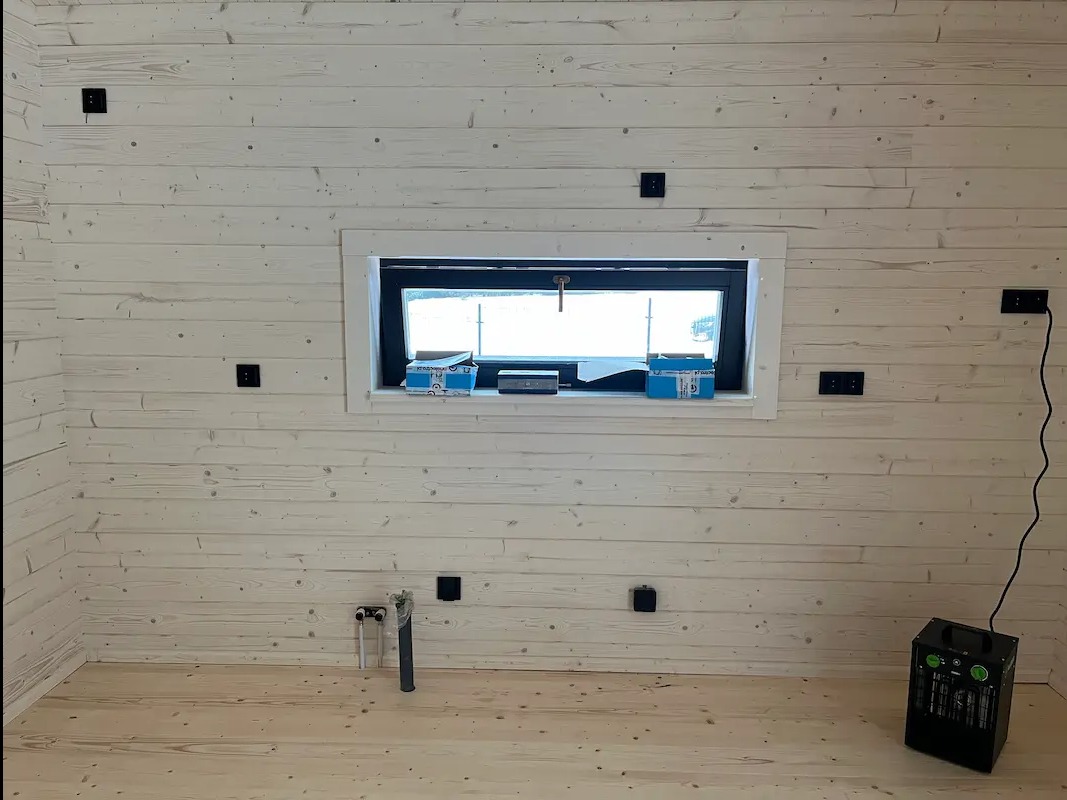 Wnętrze drewnianego domu z widocznymi puszkami elektrycznymi na ścianach, instalacją hydrauliczną i grzejnikiem elektrycznym. Przygotowanie do wykończenia.