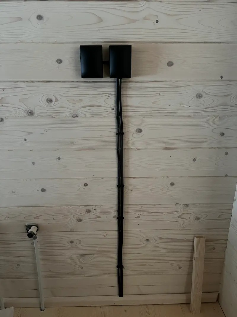 Dwa czarne, kwadratowe reflektory zamontowane na jasnej, drewnianej ścianie. Kable prowadzone pionowo w dół, przymocowane uchwytami. Widoczny fragment instalacji wodnej.