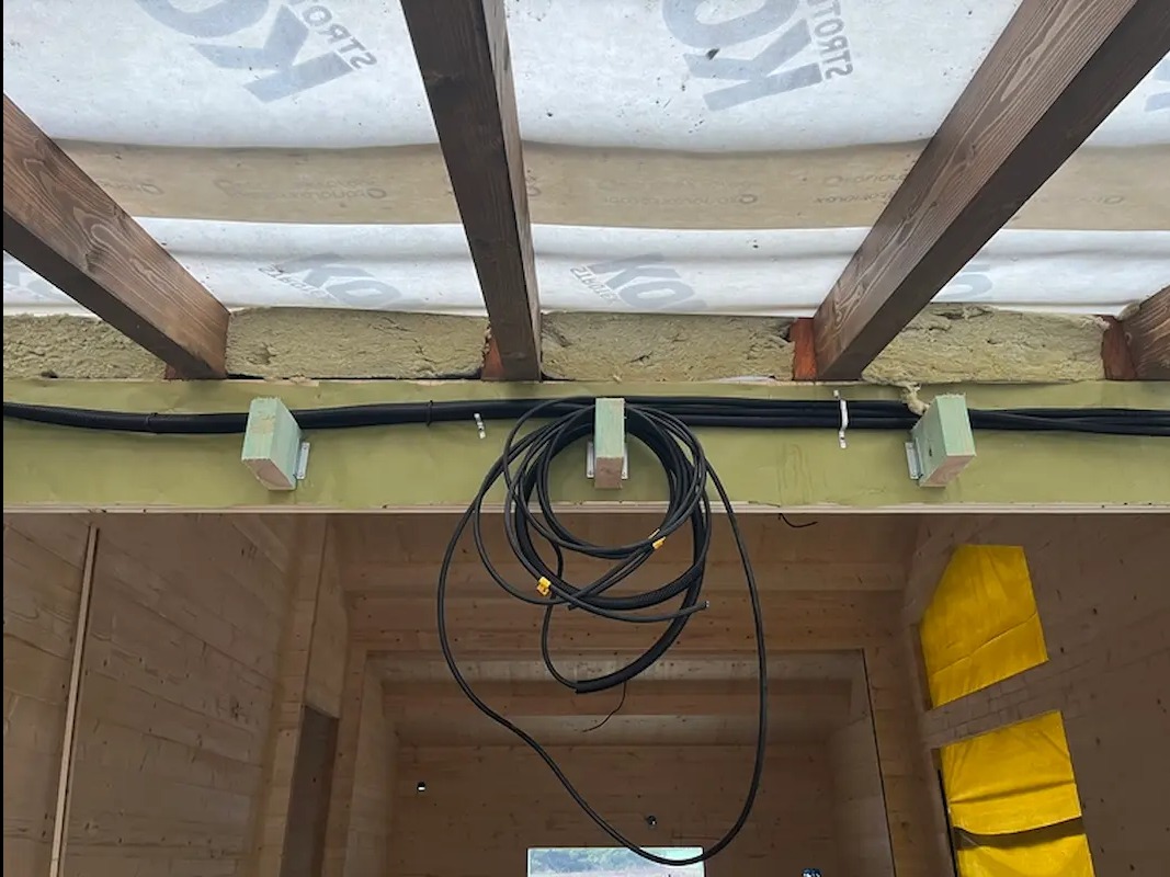 Kable elektryczne zamontowane pod sufitem w drewnianym domu, widoczne izolacja i elementy konstrukcyjne. Kabel zwisający w pętli. Ujęcie z dołu, wnętrze w trakcie budowy.