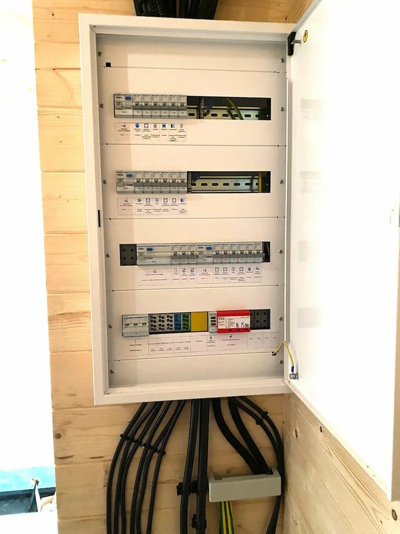 Otwarta, biała skrzynka rozdzielcza z bezpiecznikami i okablowaniem na drewnianej ścianie. Widoczne czarne kable wchodzące do skrzynki. Instalacja elektryczna w domu.