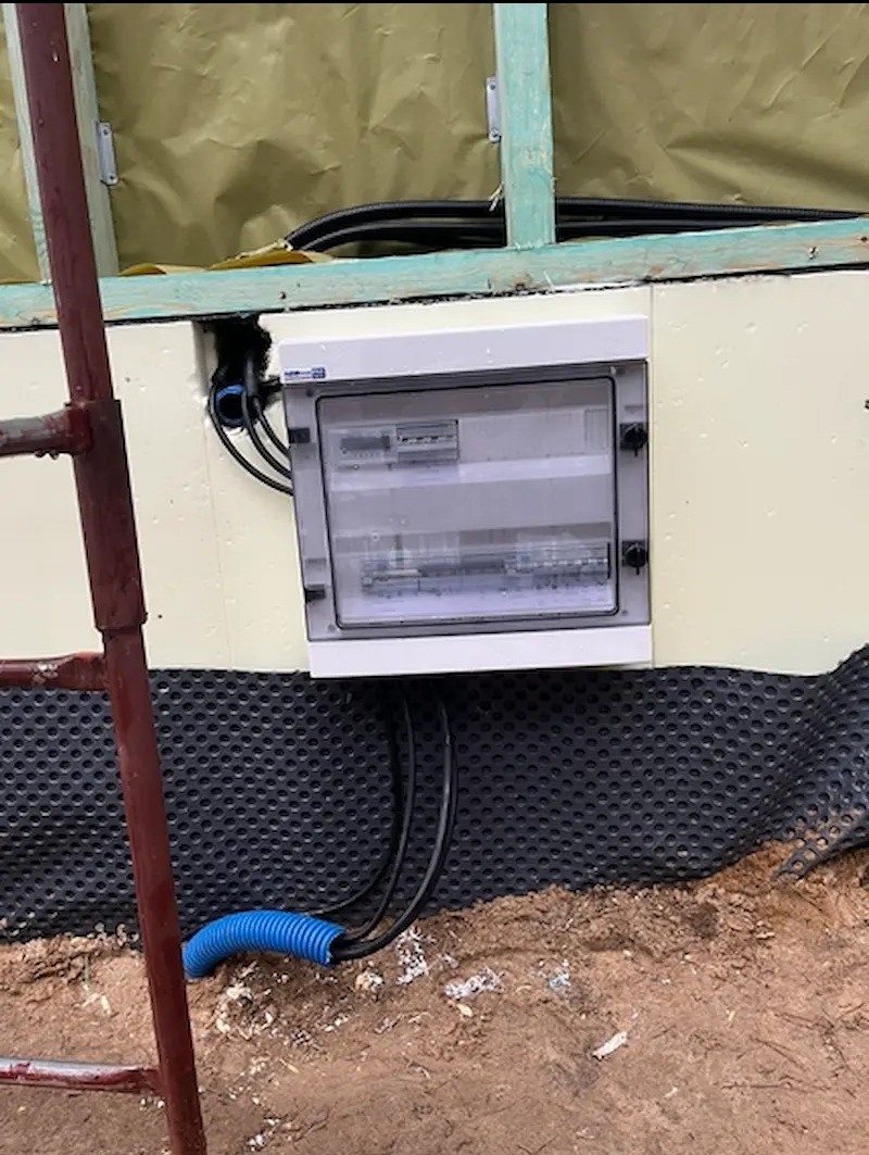 Zamontowana skrzynka elektryczna z przewodami na zewnątrz budynku w trakcie budowy, widoczne elementy konstrukcyjne i izolacja.