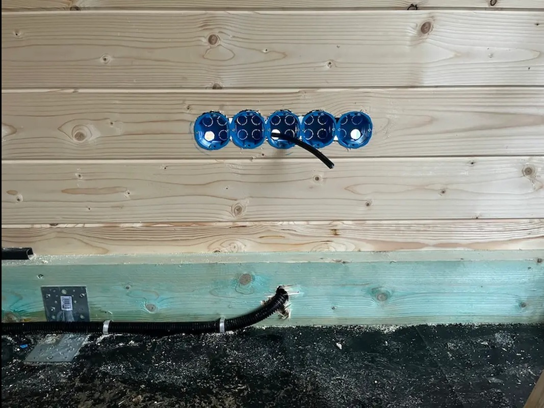 Drewniana ściana z zamontowanymi, niebieskimi puszkami elektrycznymi w rzędzie. Widoczne kable w peszlu. Surowy stan, instalacja w trakcie realizacji.