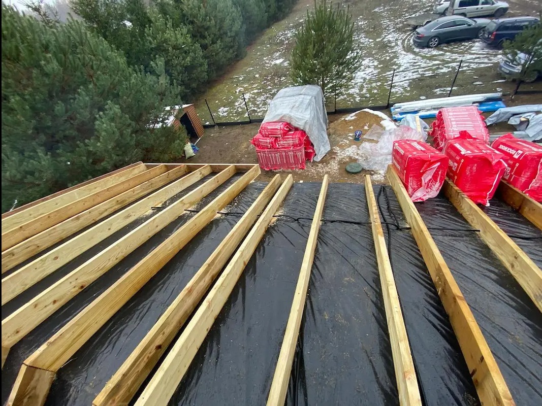 Konstrukcja dachu z drewnianymi belkami i czarną membraną, przygotowana do dalszych prac. W tle widoczne materiały izolacyjne i otoczenie z drzewami.