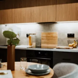 Nowoczesna kuchnia z drewnianymi i czarnymi szafkami, blatem imitującym beton, oświetleniem LED podszafkowym i okrągłym stołem z szarą zastawą w minimalistycznym stylu.