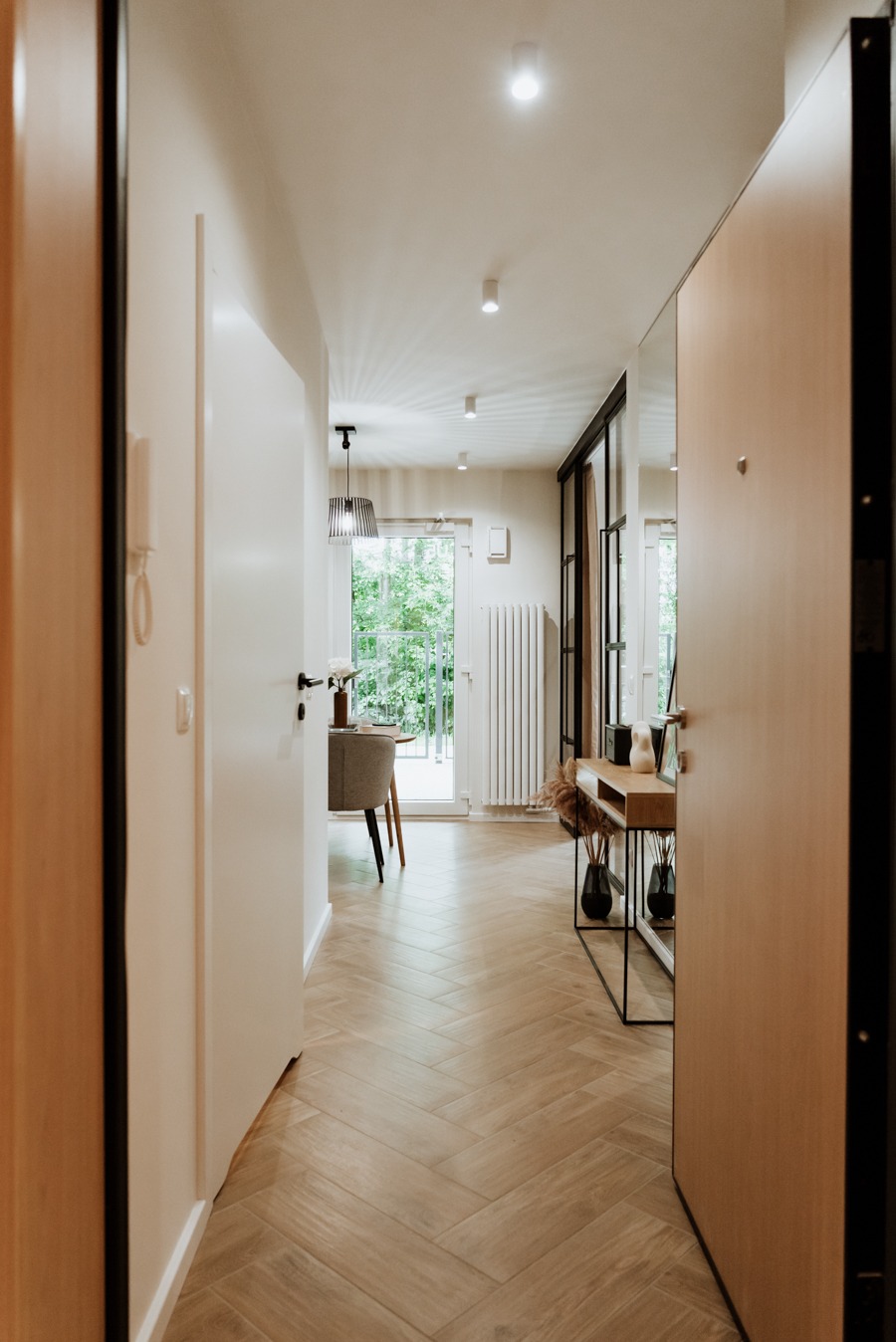 Widok na korytarz w mieszkaniu z parkietem ułożonym w jodełkę, konsolą z dekoracjami i widokiem na balkon z zielenią w tle.