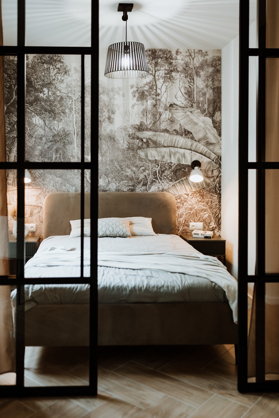 Sypialnia z łóżkiem z beżowym zagłówkiem, pościelą w odcieniach szarości i tapetą z motywem dżungli, widoczna przez czarne, przeszklone drzwi przesuwne.