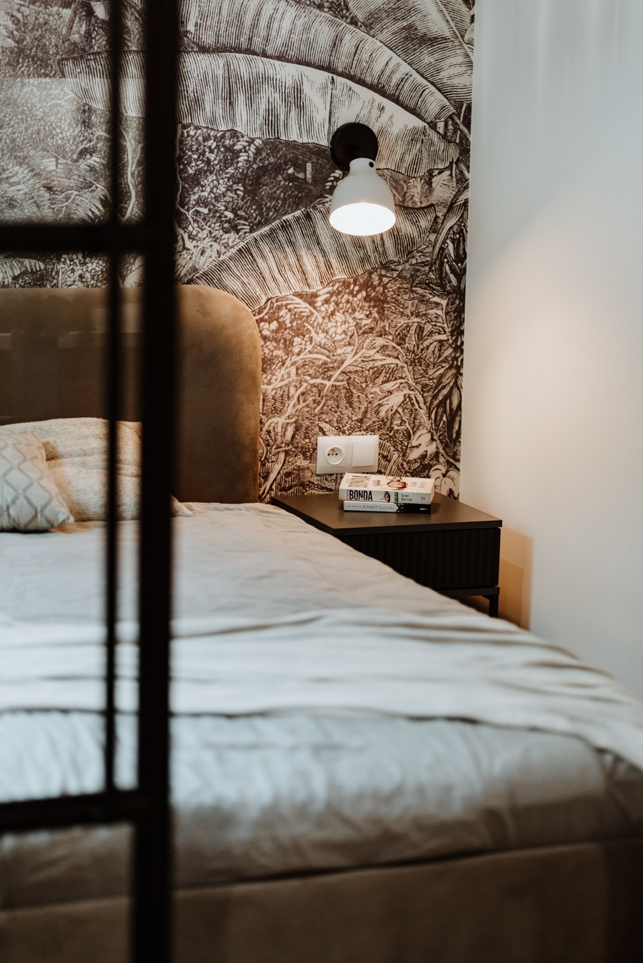 Sypialnia z łóżkiem z szarym zagłówkiem, szafką nocną z lampką i książkami, oraz tapetą w motywy roślinne, widziana przez metalową ramę.