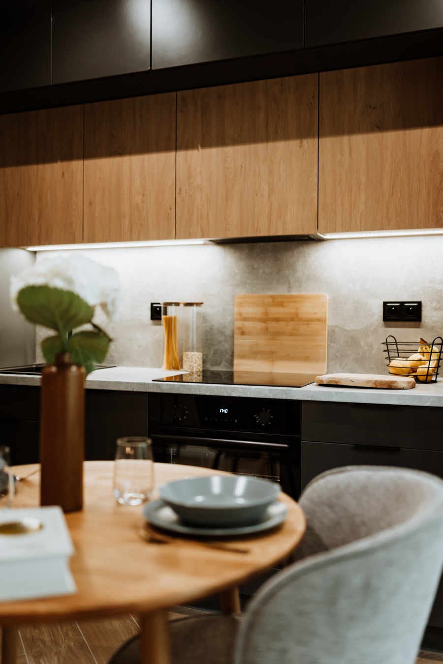 Nowoczesna kuchnia z drewnianymi i czarnymi szafkami, blatem imitującym beton, oświetleniem LED podszafkowym i okrągłym stołem z szarą zastawą w minimalistycznym stylu.