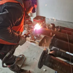 Spawacz w trakcie pracy nad rdzewiejącymi rurami z kołnierzami, iskry i intensywne światło spawania, wnętrze w trakcie remontu, widoczne narzędzia i elementy instalacji.