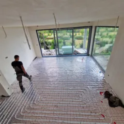 Instalacja ogrzewania podłogowego w nowym domu z dużymi oknami. Widoczny pracownik w trakcie układania rur na folii izolacyjnej. Zastosowano białe rury i czerwoną siatkę montażową.