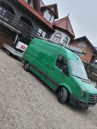 Zielony samochód dostawczy z drabiną na dachu zaparkowany na żwirowym podjeździe przed domem, z przyczepą i mini koparką w tle.