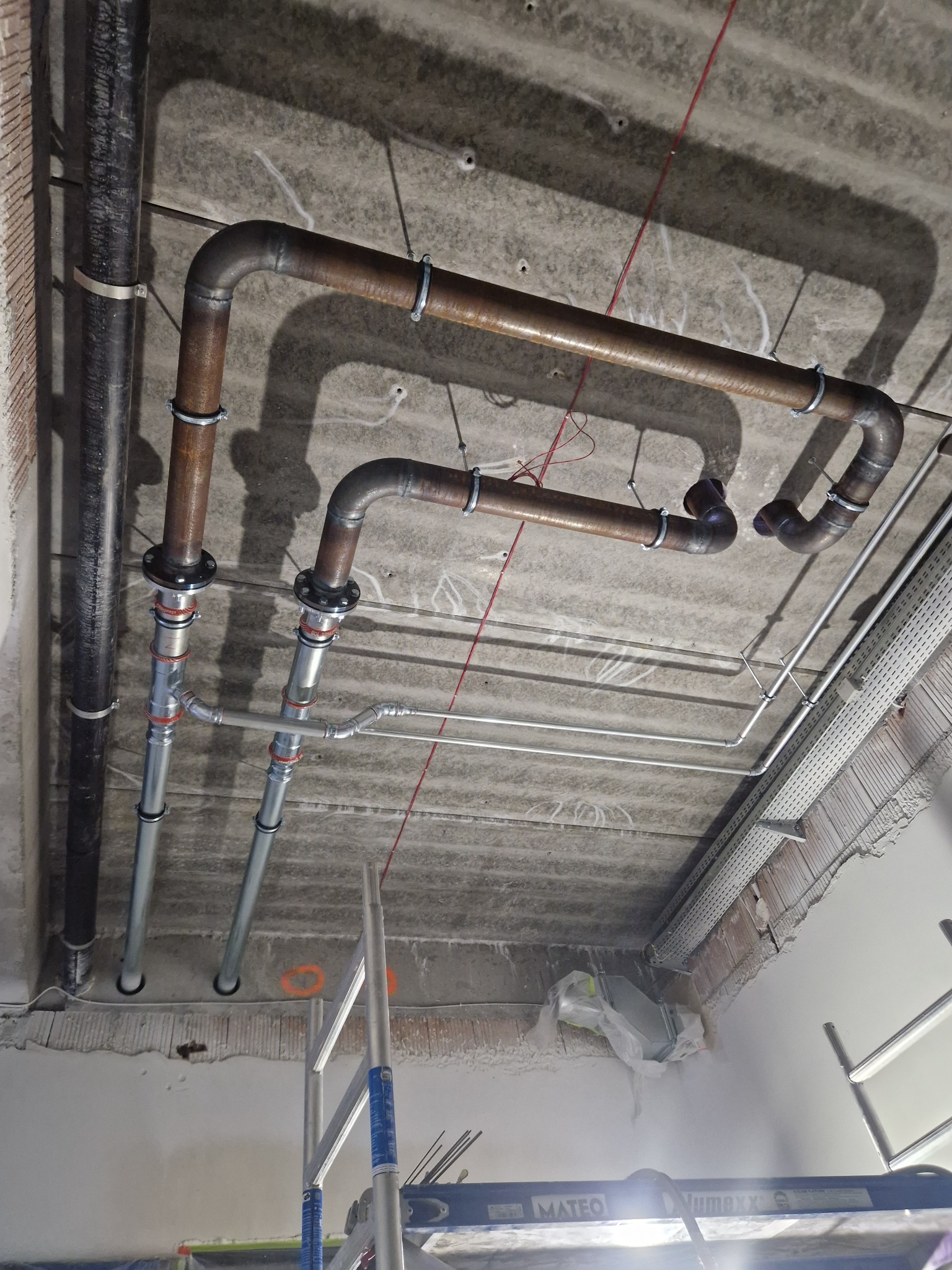 Różne rury pod stropem, widoczne mocowania i kable. Prace instalacyjne w Myślenicach, widok z dołu na sufit w trakcie montażu instalacji.