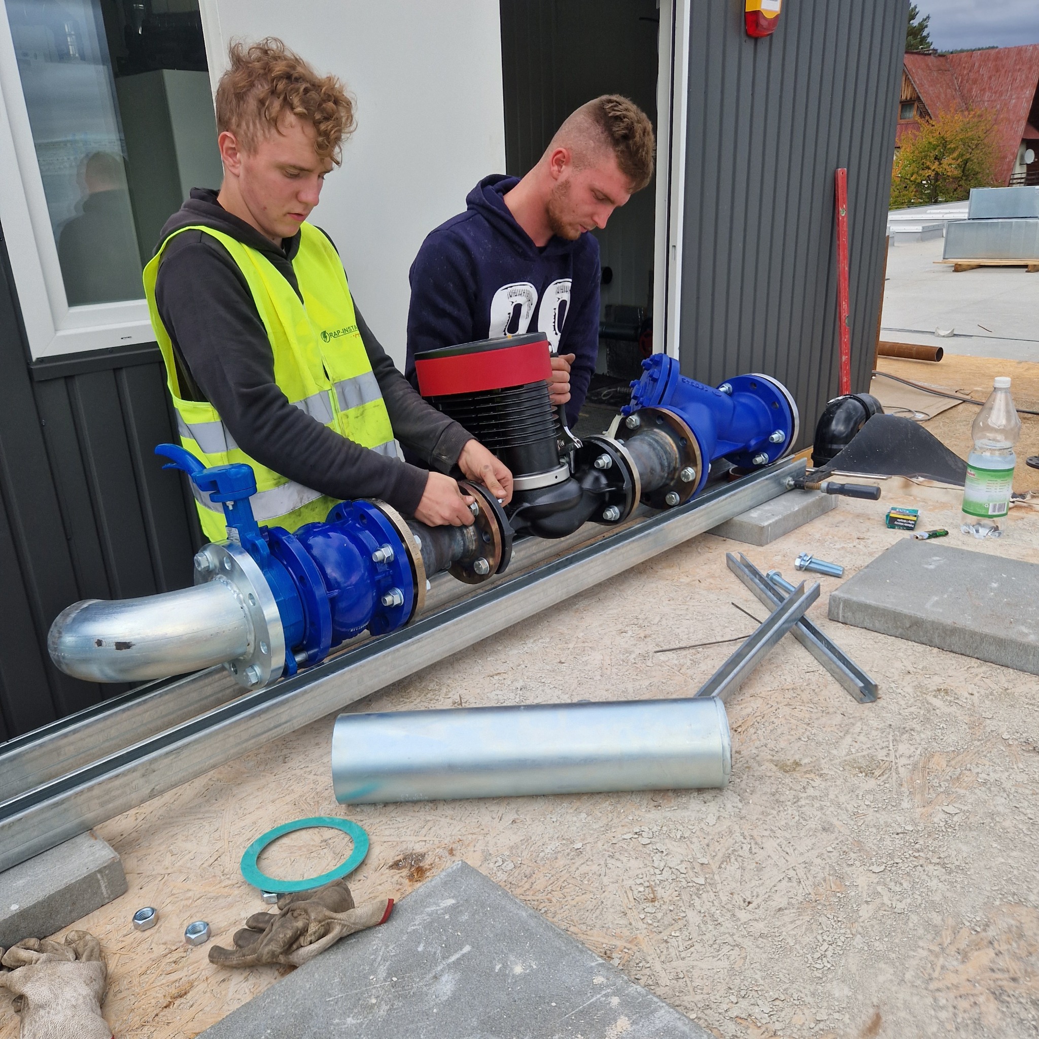 Dwóch monterów łączy niebieskie rury z czarnym urządzeniem na metalowej konstrukcji, na zewnątrz budynku. Widoczne narzędzia i elementy montażowe.