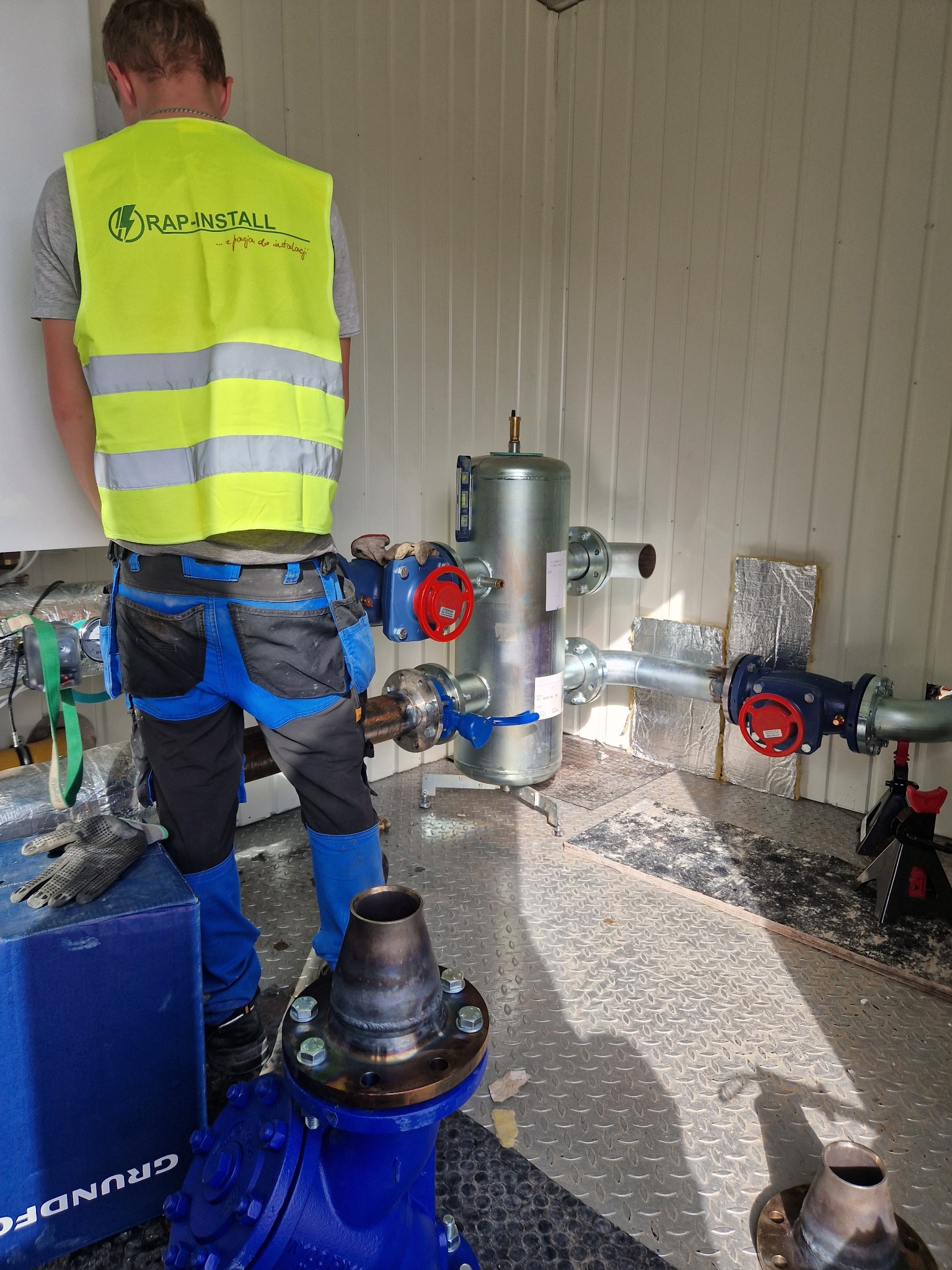 Instalator w kamizelce RAP-INSTALL montuje srebrny zbiornik z rurami i czerwonymi zaworami w pomieszczeniu o metalowej podłodze.