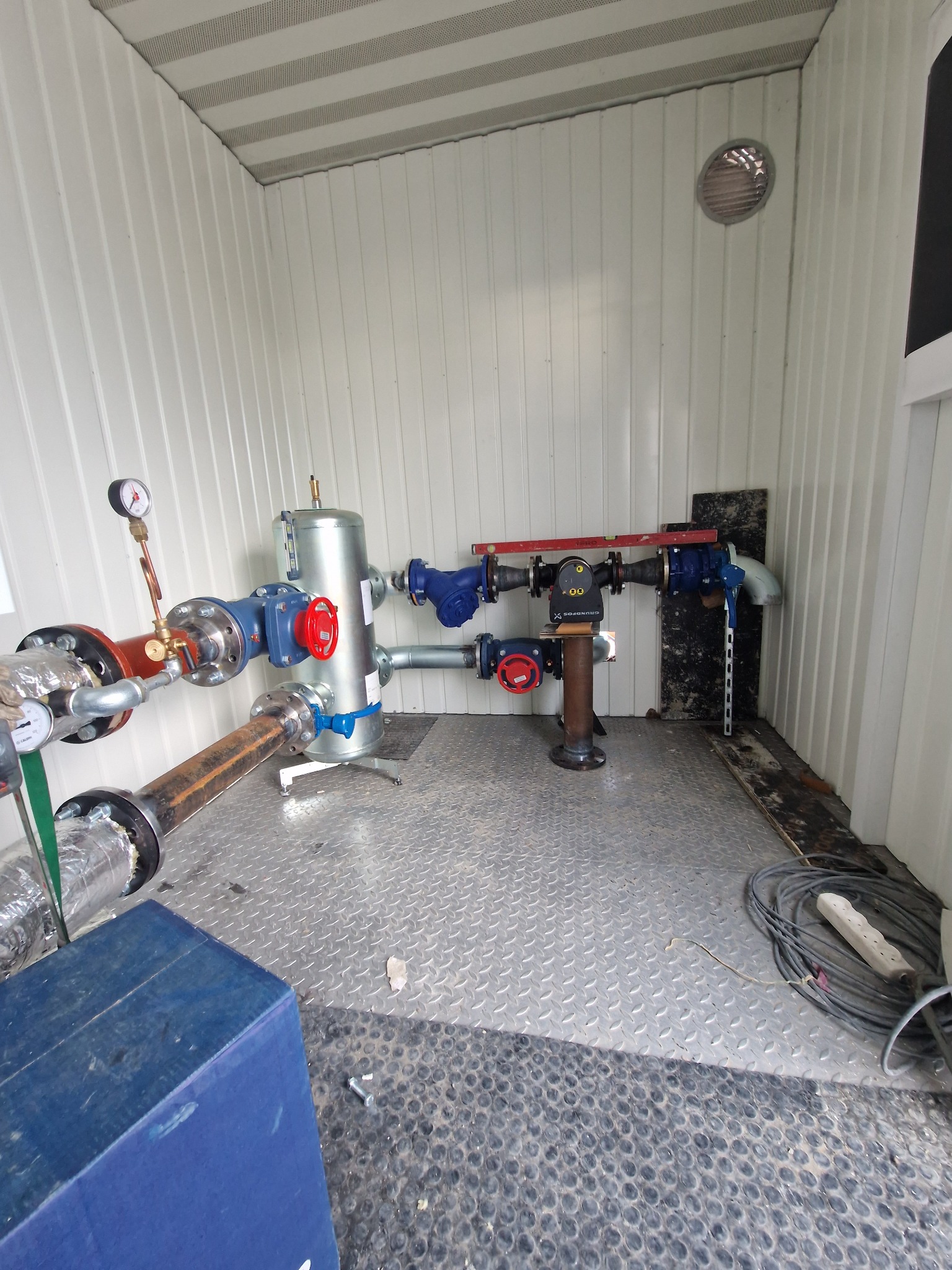 Węzeł cieplny w kontenerze: metalowy zbiornik, rury z zaworami (czerwone, niebieskie), manometry. Podłoga blaszana, ściany z blachy trapezowej. Widoczna poziomica.