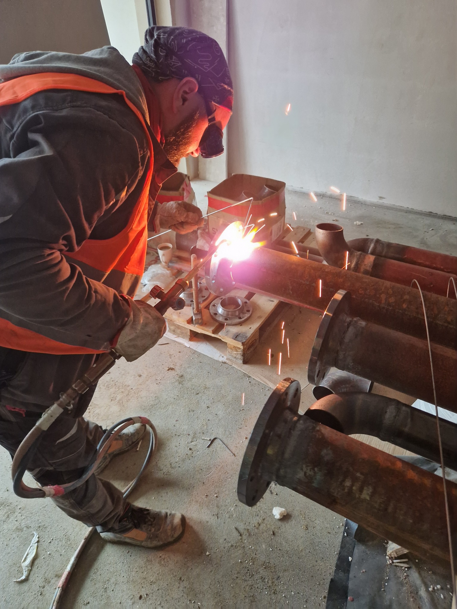 Spawacz w trakcie pracy nad rdzewiejącymi rurami z kołnierzami, iskry i intensywne światło spawania, wnętrze w trakcie remontu, widoczne narzędzia i elementy instalacji.