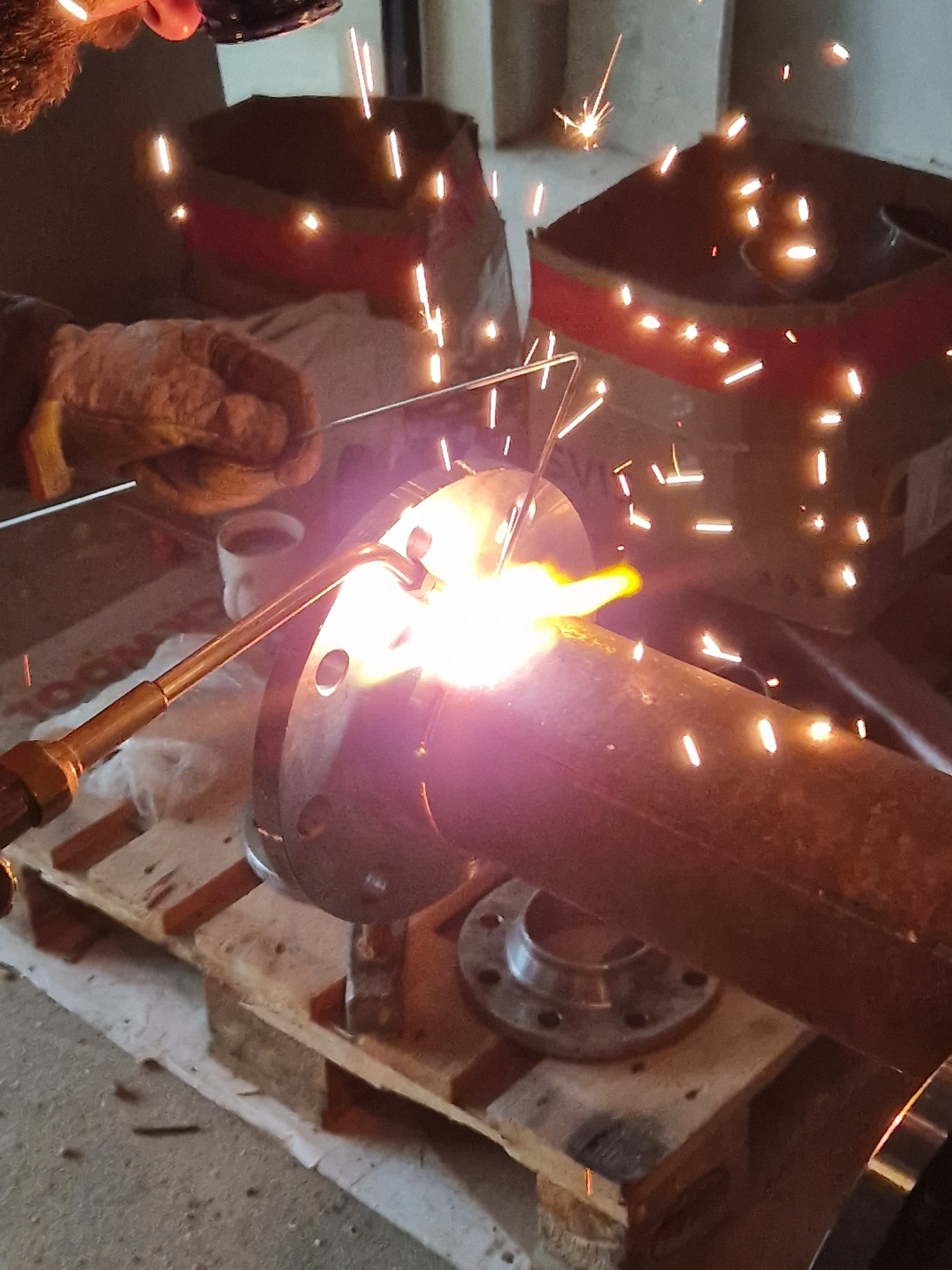 Spawanie rury z kołnierzem na palecie, iskry rozświetlają otoczenie. Detal z bliska ukazuje precyzję łączenia elementów instalacji.