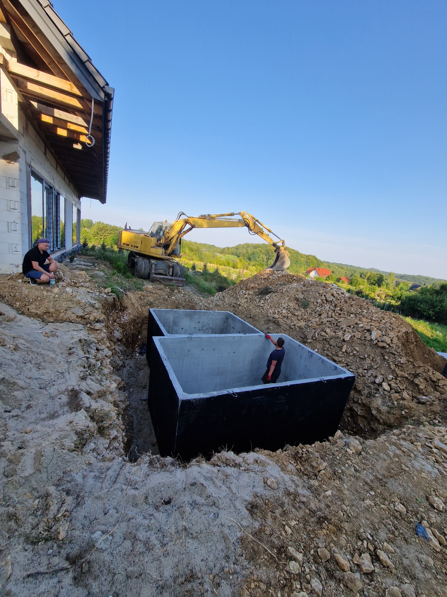 Montaż betonowego zbiornika na deszczówkę w wykopie. Koparka i pracownicy na budowie domu jednorodzinnego. Widok na wzgórza w tle.