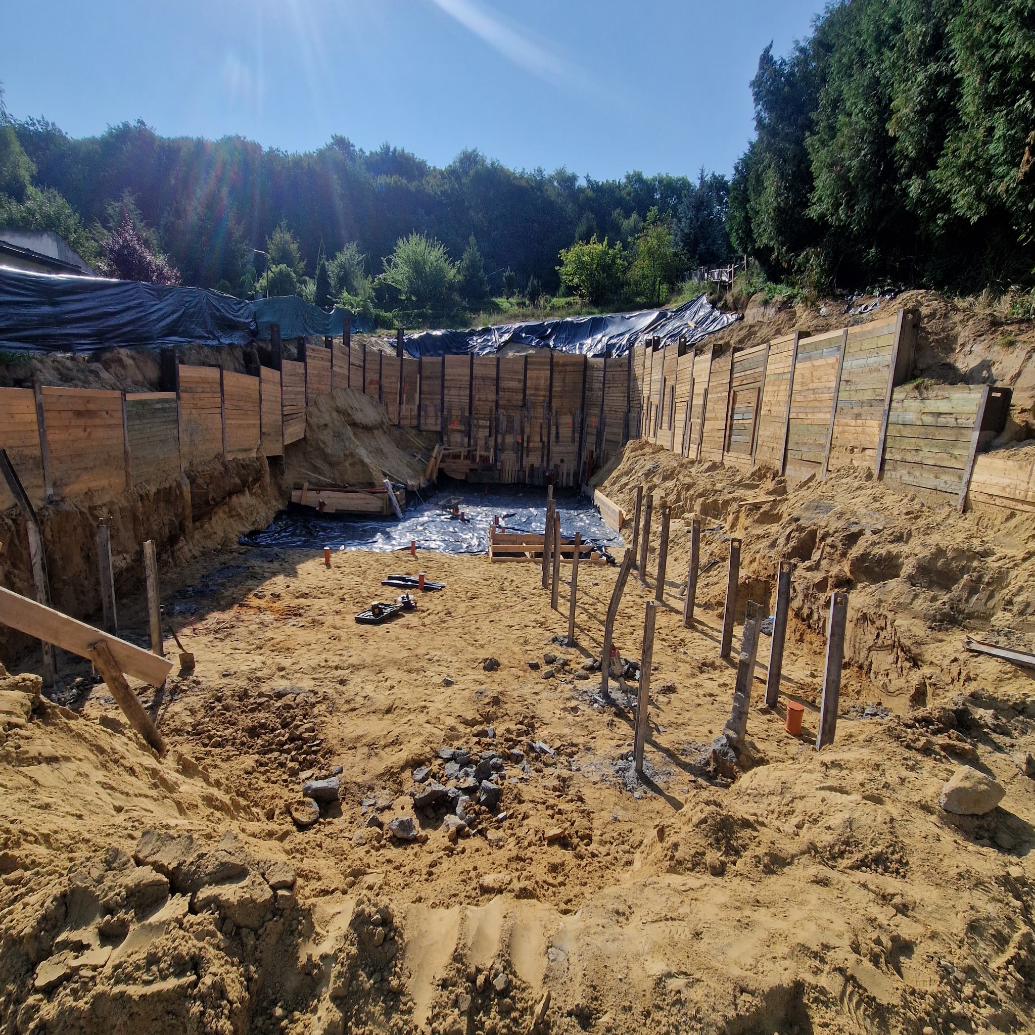 Wykop pod budowę domu zabezpieczony drewnianą konstrukcją i folią. Widoczne narzędzia i elementy instalacji. Tło stanowią drzewa i niebo.