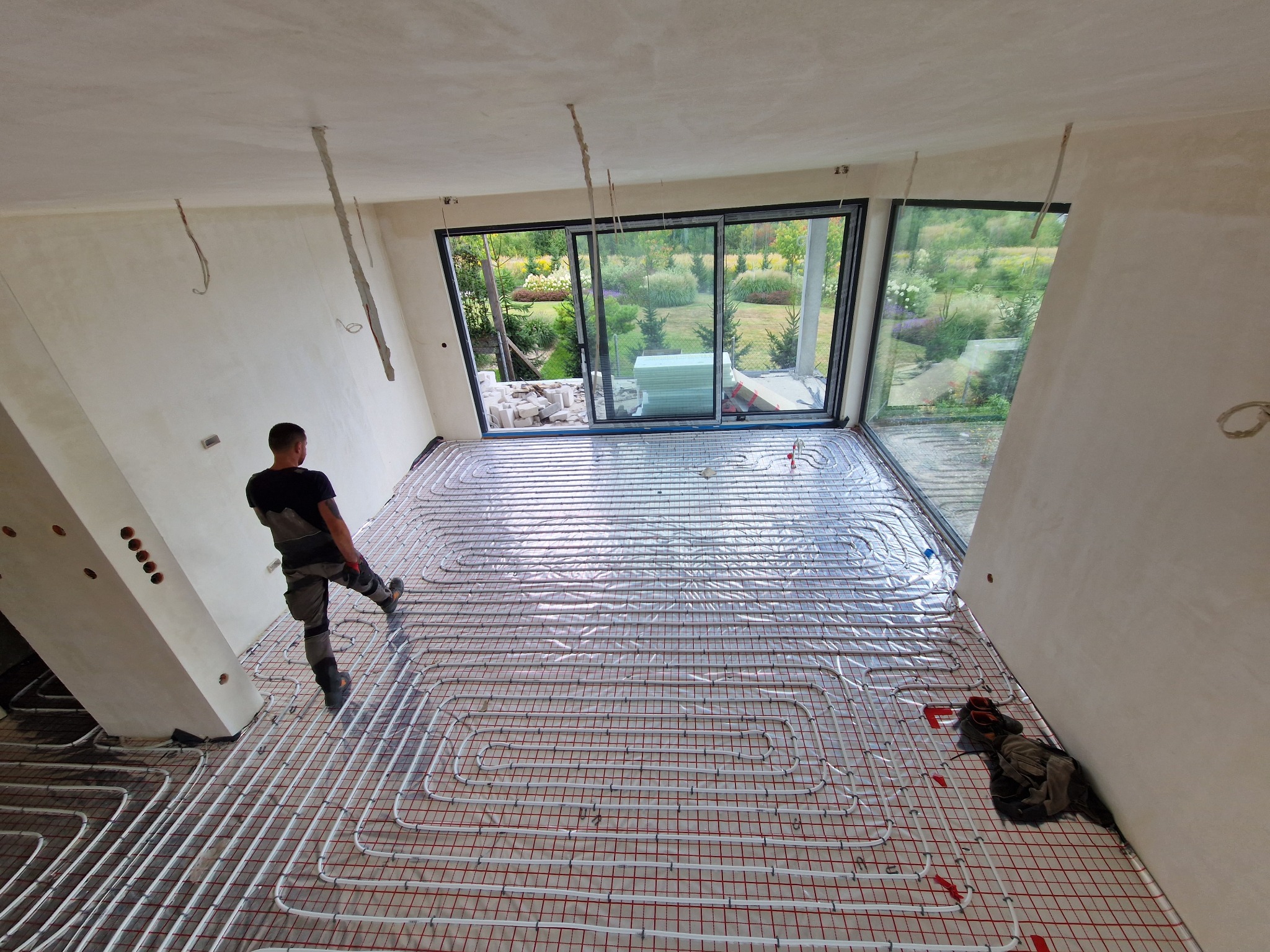 Instalacja ogrzewania podłogowego w nowym domu z dużymi oknami. Widoczny pracownik w trakcie układania rur na folii izolacyjnej. Zastosowano białe rury i czerwoną siatkę montażową.
