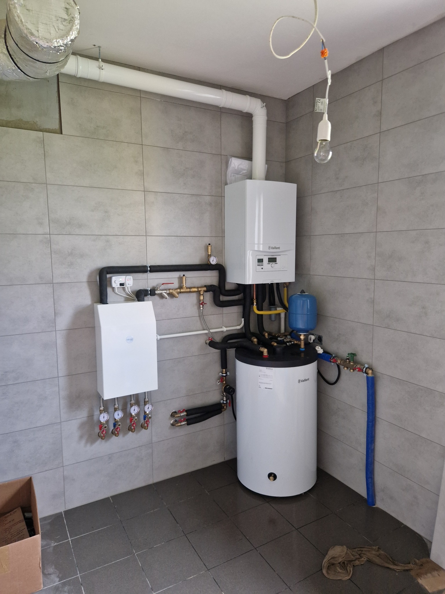 Zestaw urządzeń grzewczych: biały piec gazowy Vaillant, zbiornik, naczynie przeponowe i rozdzielacz w pomieszczeniu wykończonym szarą płytką. Widoczna instalacja i rury.