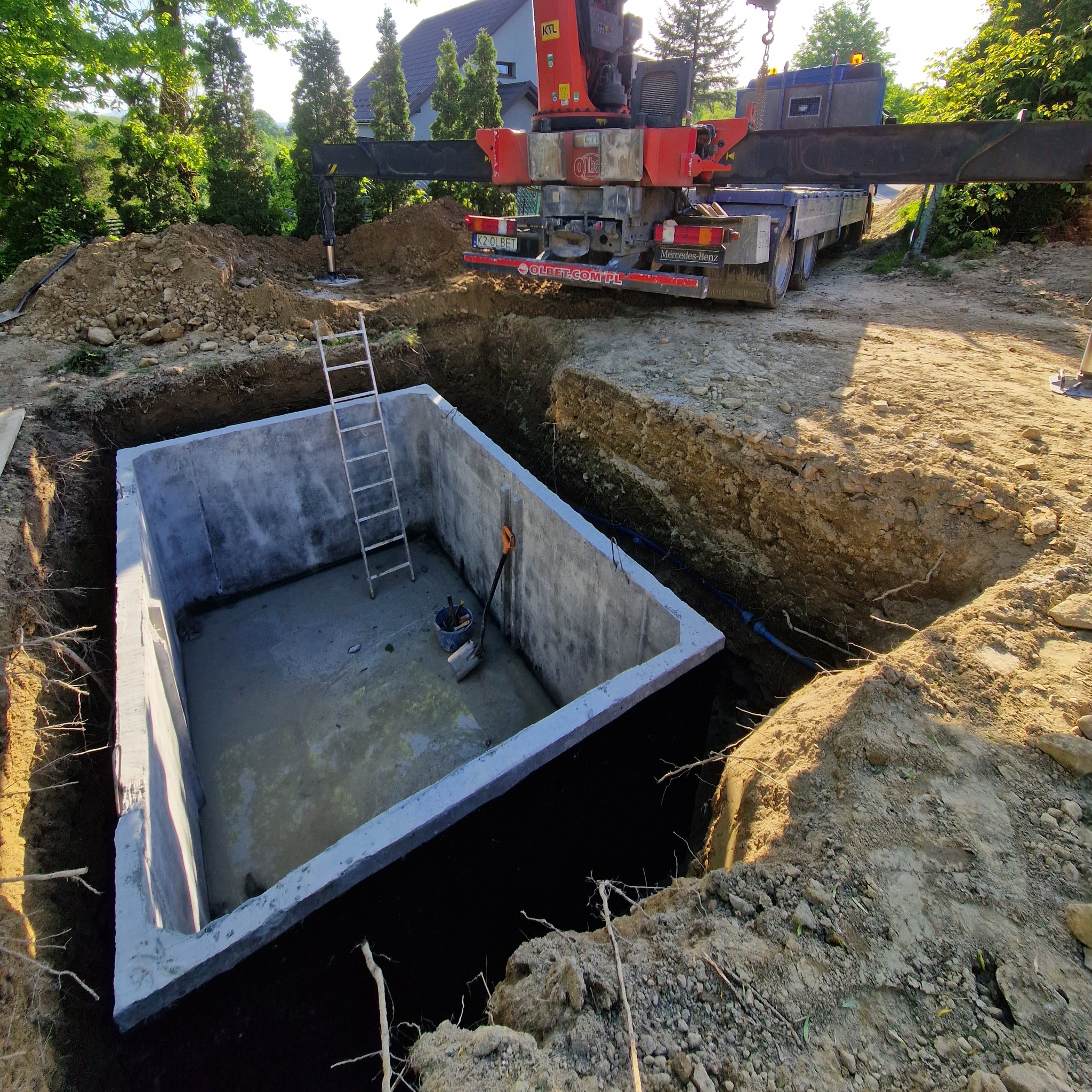 Betonowy zbiornik w wykopie, z drabiną i narzędziami w środku, wspierany przez dźwig. Widoczne ślady prac ziemnych i niebieski przewód. Słoneczny dzień.