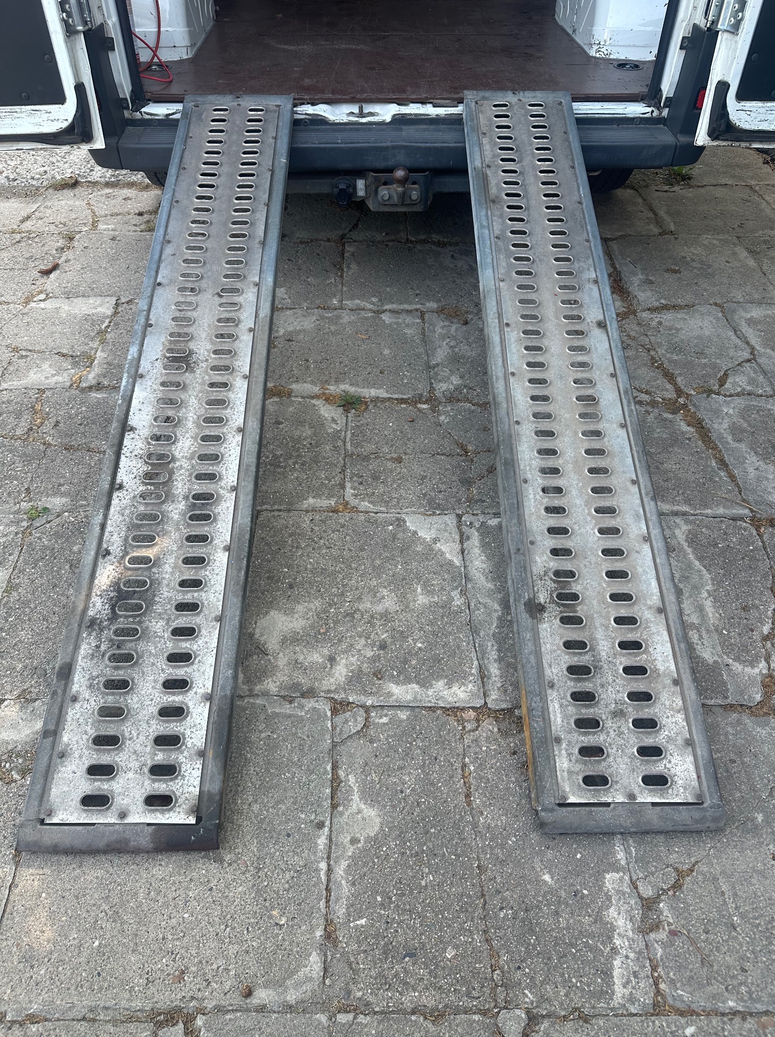 Rampa załadunkowa z perforowanej stali, wysunięta z bagażnika busa na chodnik z betonowej kostki. Ułatwienie transportu towarów podczas przeprowadzek.
