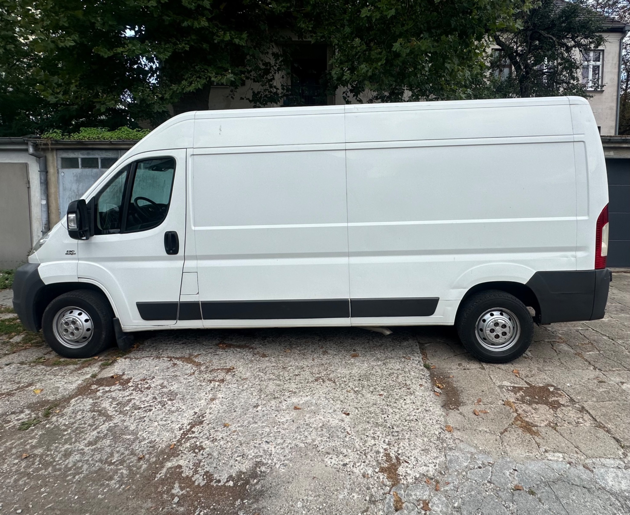 Biały samochód dostawczy Fiat Ducato zaparkowany na zewnątrz, widok z boku, zarys budynku w tle, na zewnątrz, w dzień.