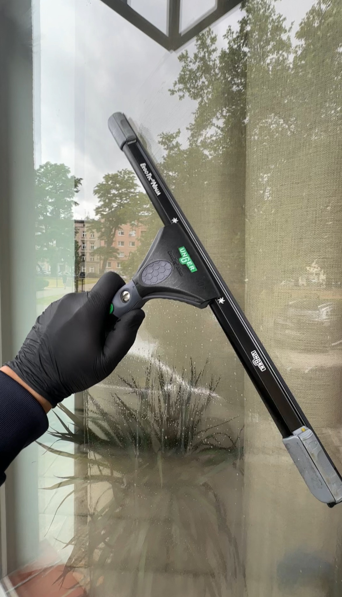 Mycie okna od wewnątrz za pomocą ściągaczki Unger ErgoTec Ninja. Widoczna ręka w czarnej rękawicy i budynek za oknem. Ślady wody na szybie.