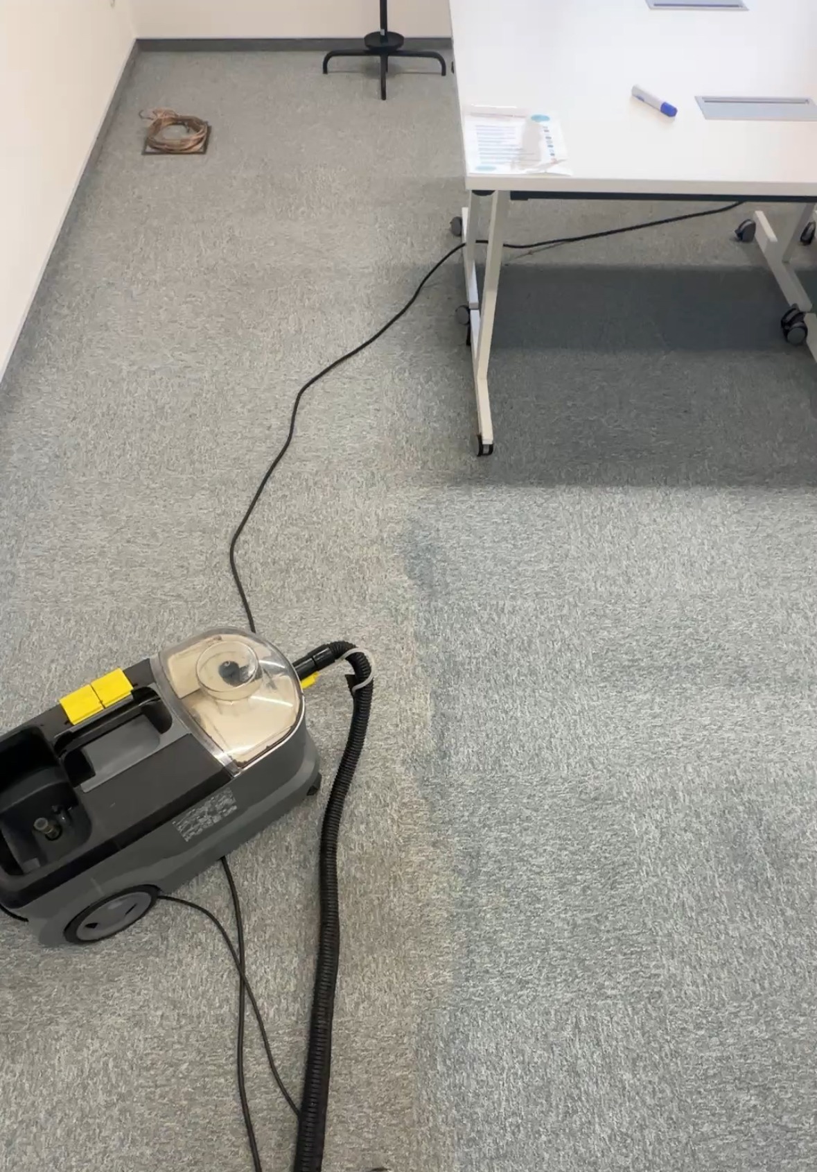 Szaro-żółty odkurzacz piorący na tle wyczyszczonej i brudnej wykładziny w biurze. Widoczny fragment biurka i krzesła. Prezentacja efektu czyszczenia.