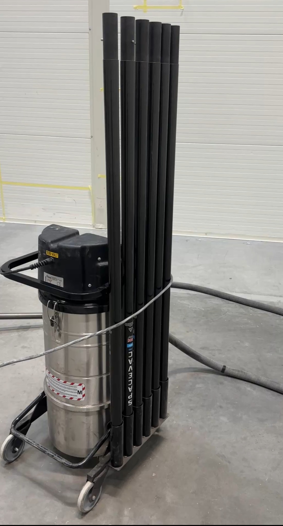 Profesjonalny odkurzacz przemysłowy z zestawem rur ssących na wózku, gotowy do użycia w przestronnym, jasnym pomieszczeniu po remoncie. Widoczne ślady po pracach.