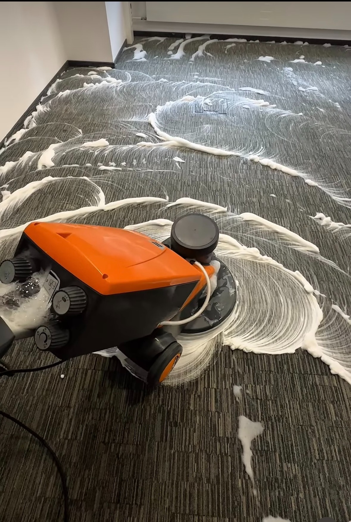 Profesjonalne pranie wykładziny dywanowej z użyciem specjalistycznej maszyny. Widoczne ślady piany czyszczącej na szarej wykładzinie.