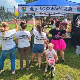 Aparatka- Animator zabaw dziecięcych. - Grupa osób w koszulkach z napisem 'Sołectwo Koszwały' i kobieta w czarnej koszulce z logo aparatu i różowej tiulowej spódnicy, stojąca pod namiotem z napisem 'Koszwały', z dzieckiem na rowerku...