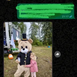 Dziewczynka w różowej kurtce przytula się do animatora przebranego za misia w czarnym fraku i cylindrze na tle zielonego trawnika i sceny.