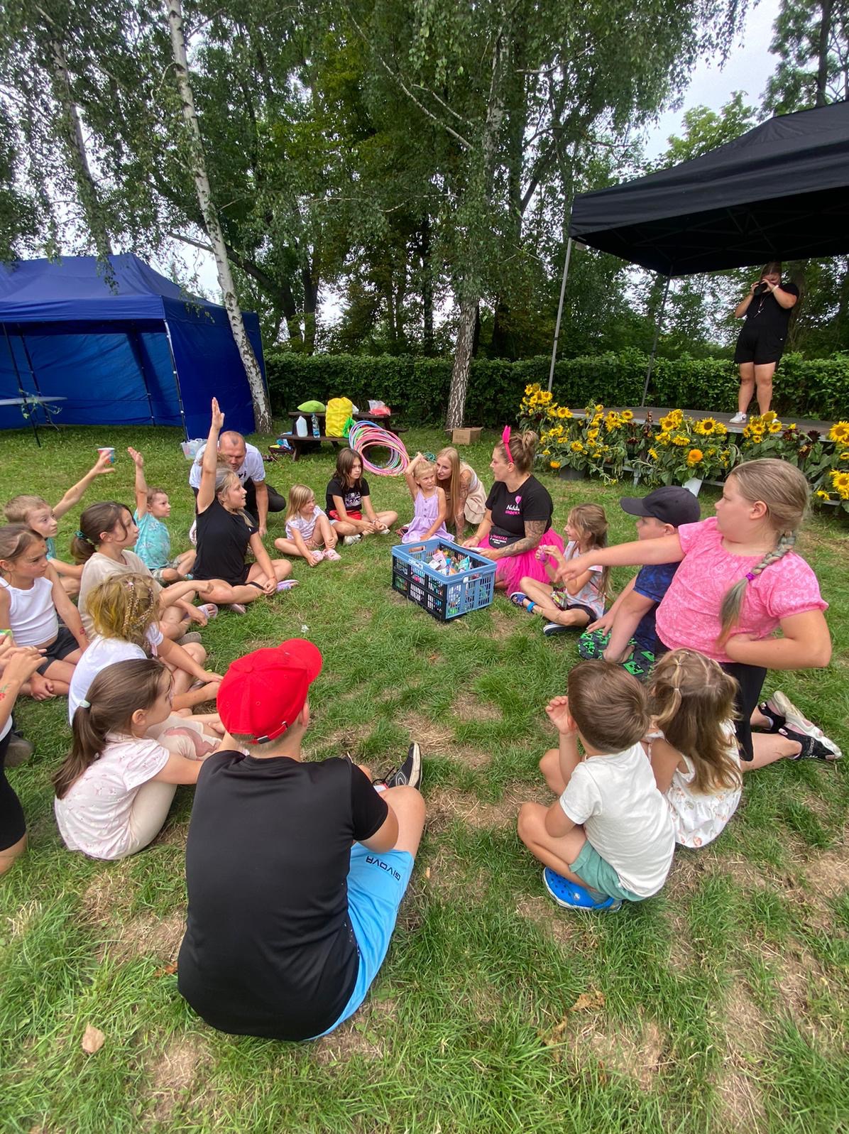 Animator prowadzi zajęcia z dziećmi na trawie w Gdańsku, w tle scena z osobą mówiącą do mikrofonu i namiot.