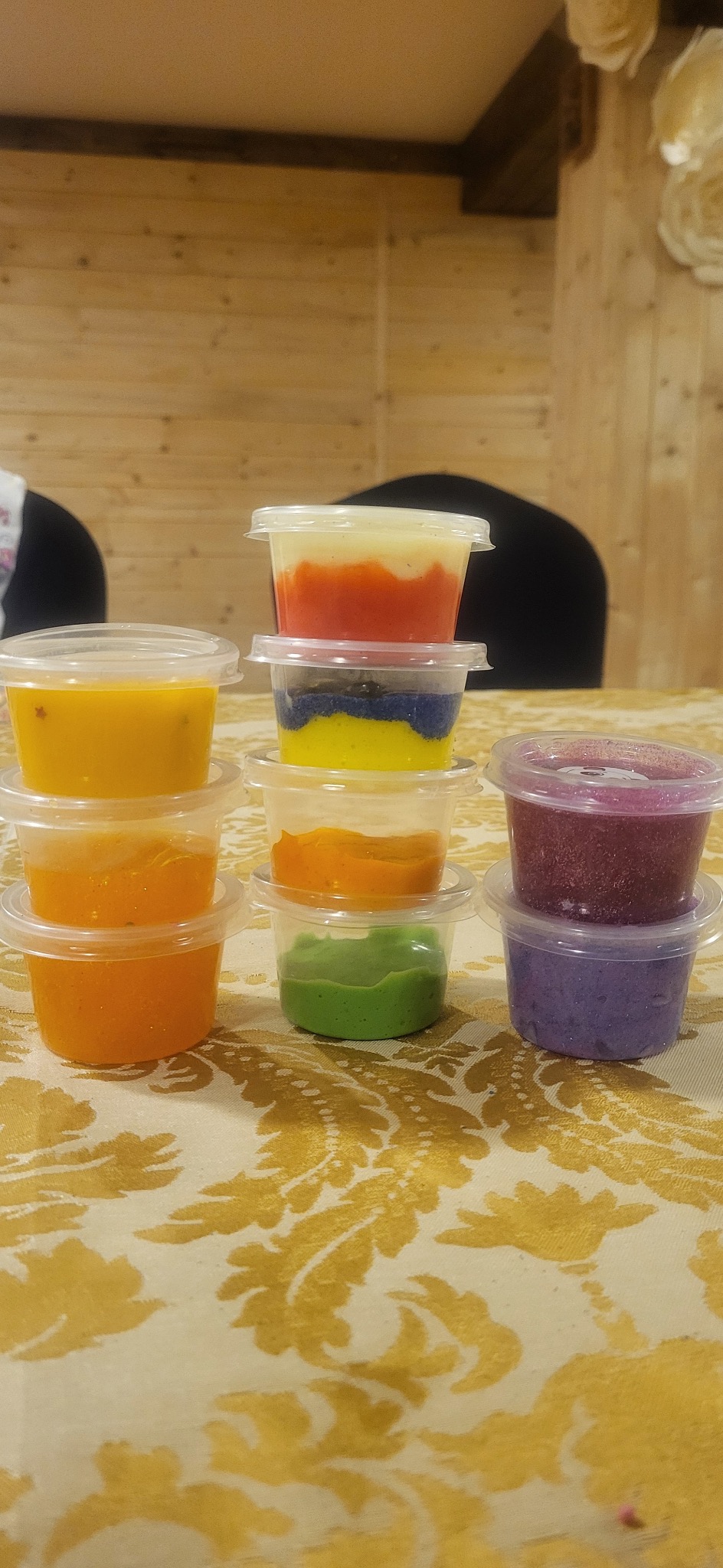 Wieża z pojemników z kolorową masą plastyczną na stole z wzorzystym obrusem, widoczne kolory: żółty, pomarańczowy, zielony, niebieski, czerwony, fioletowy i biały.