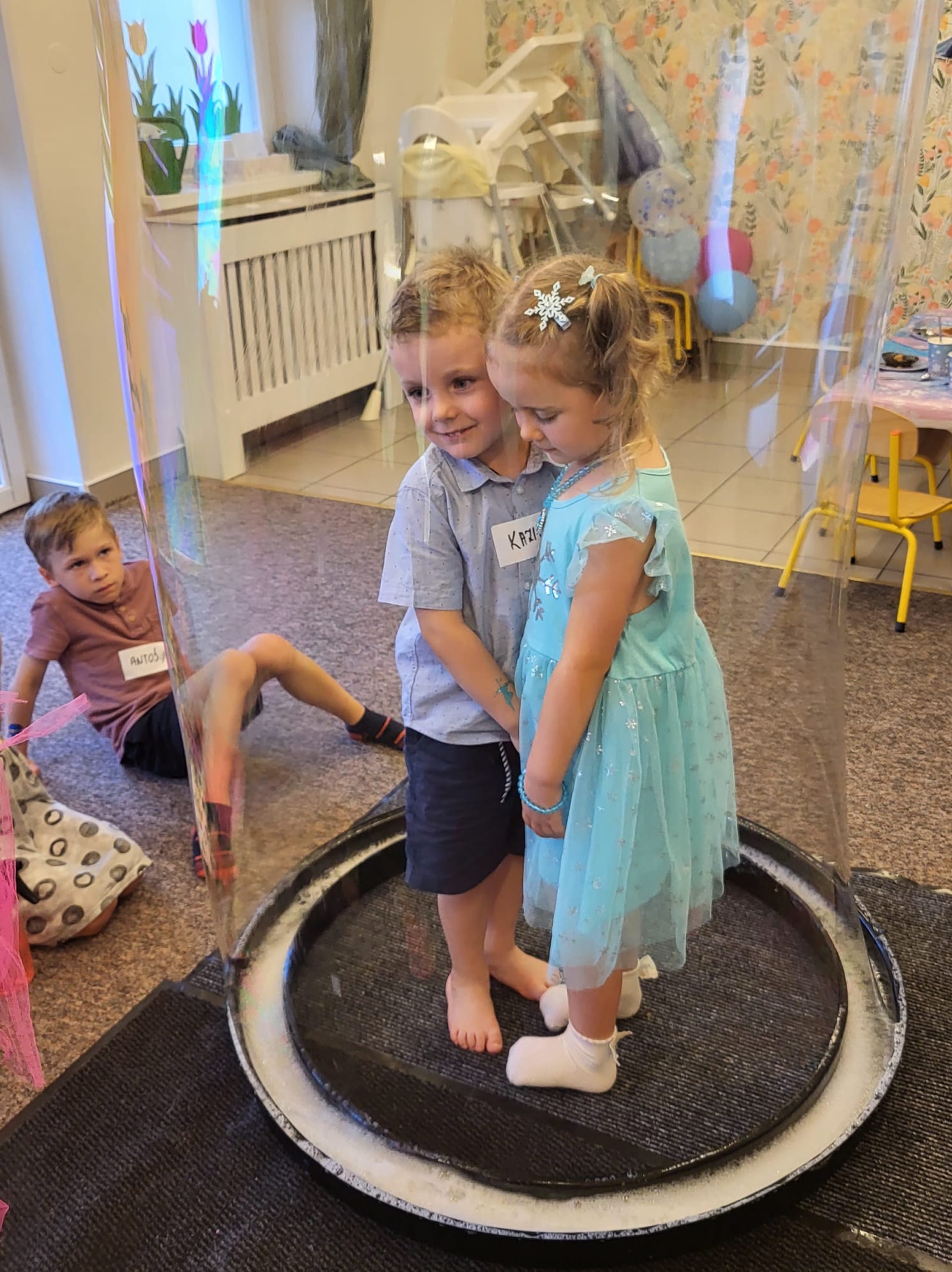 Dwoje dzieci wewnątrz dużej bańki mydlanej podczas animacji. Chłopiec w szarej koszuli obejmuje dziewczynkę w błękitnej sukience. Trzecie dziecko obserwuje z boku.