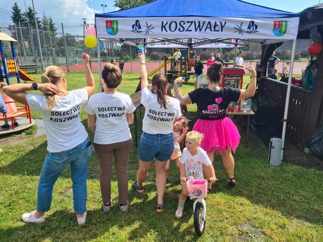 Grupa osób w koszulkach z napisem 'Sołectwo Koszwały' i kobieta w czarnej koszulce z logo aparatu i różowej tiulowej spódnicy, stojąca pod namiotem z napisem 'Koszwały', z dzieckiem na rowerku...