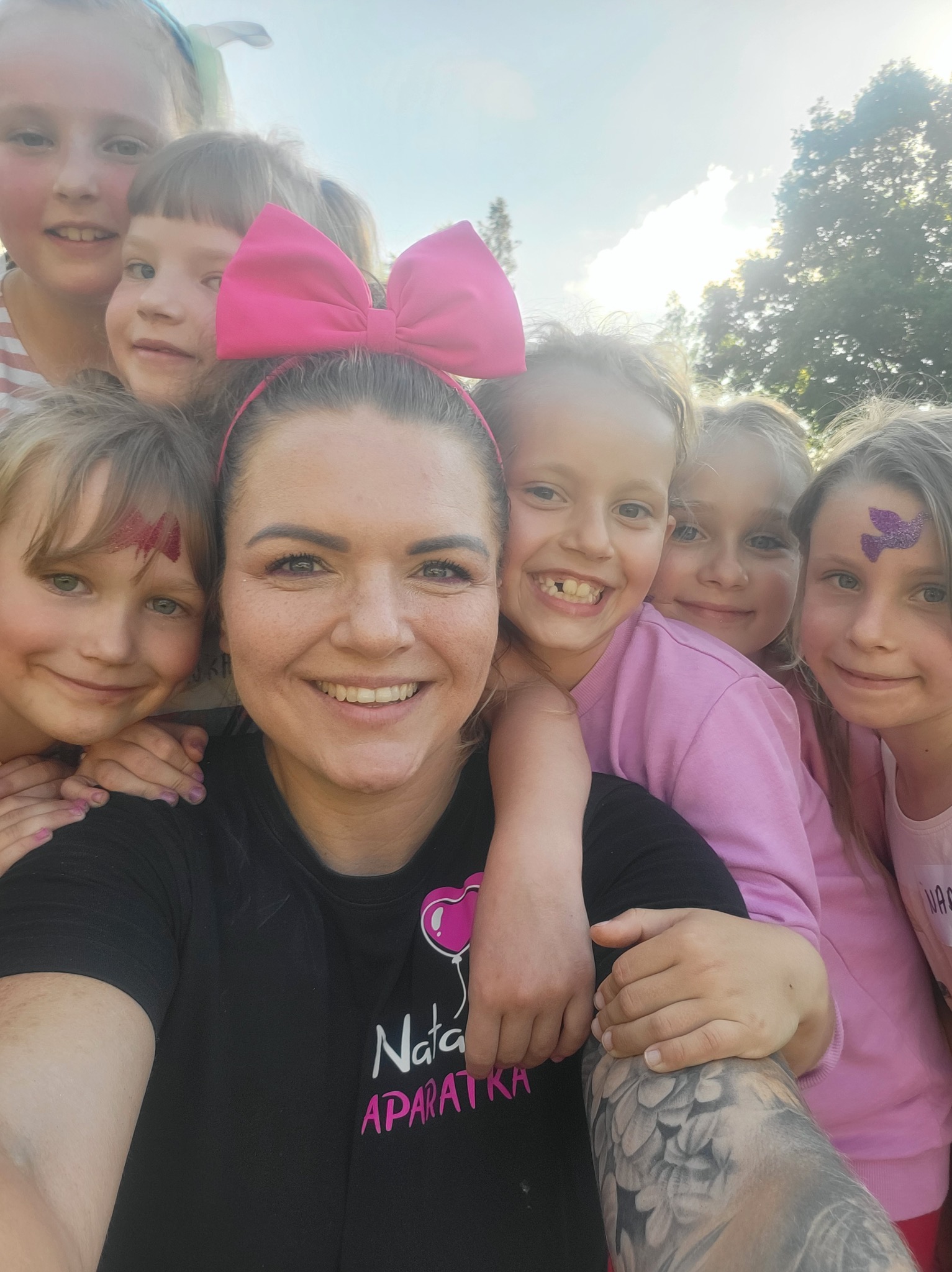 Animator z dziećmi podczas imprezy, dziewczynki z pomalowanymi twarzami i różową kokardą na głowie, widoczny tatuaż na ramieniu animatora.