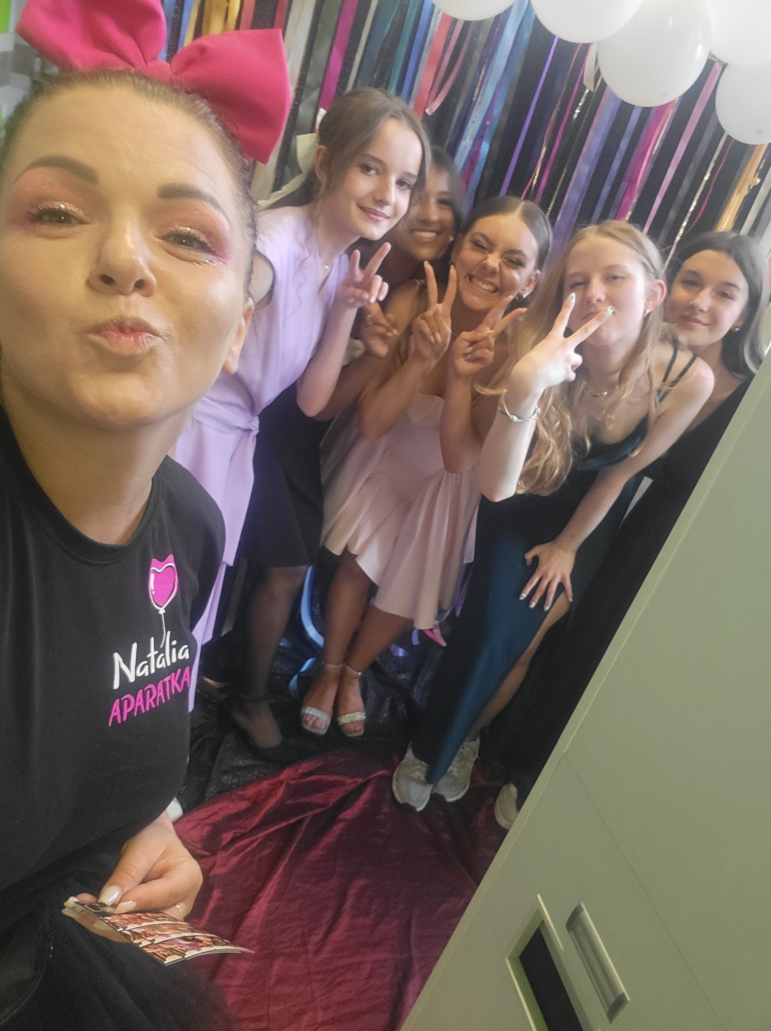 Zdjęcie selfie z grupą nastolatek pozujących z gestem V przed tłem z balonów i wstążek, z operatorką fotobudki trzymającą wydrukowane zdjęcia.