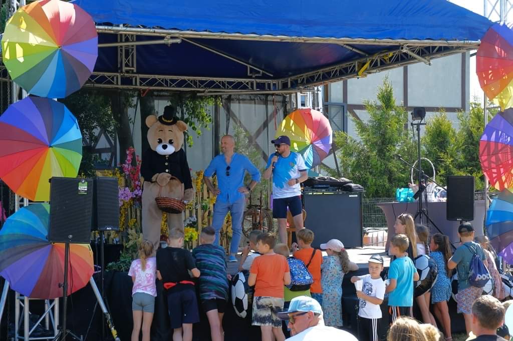 Scena plenerowa z animatorem w kostiumie misia w meloniku, dwoma mężczyznami z mikrofonami i grupą dzieci oglądających przedstawienie, udekorowana kolorowymi parasolkami.
