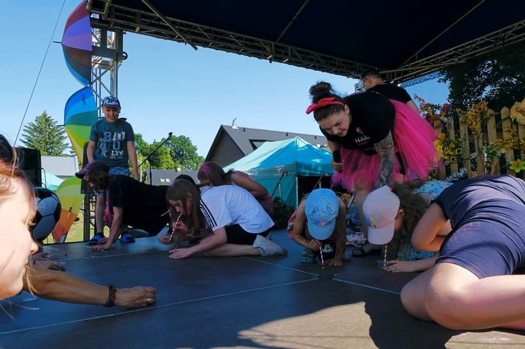 Dzieci na scenie podczas zabawy z piciem przez słomkę, nadzorowane przez dorosłych, tęczowy parasol w tle.