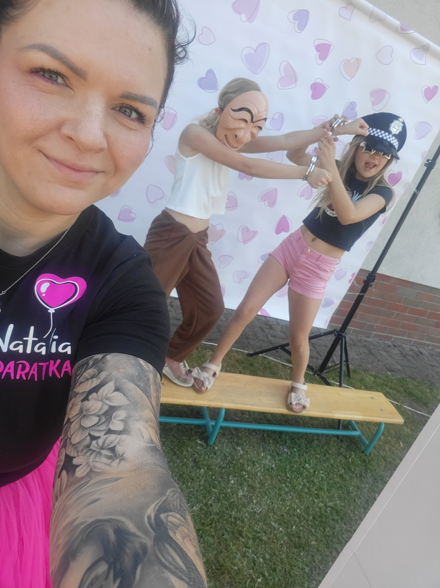 Kobieta robiąca selfie na tle scenki z dwójką dzieci: jedno w masce, drugie w czapce policjanta z kajdankami na rękach, tło z motywem serc.