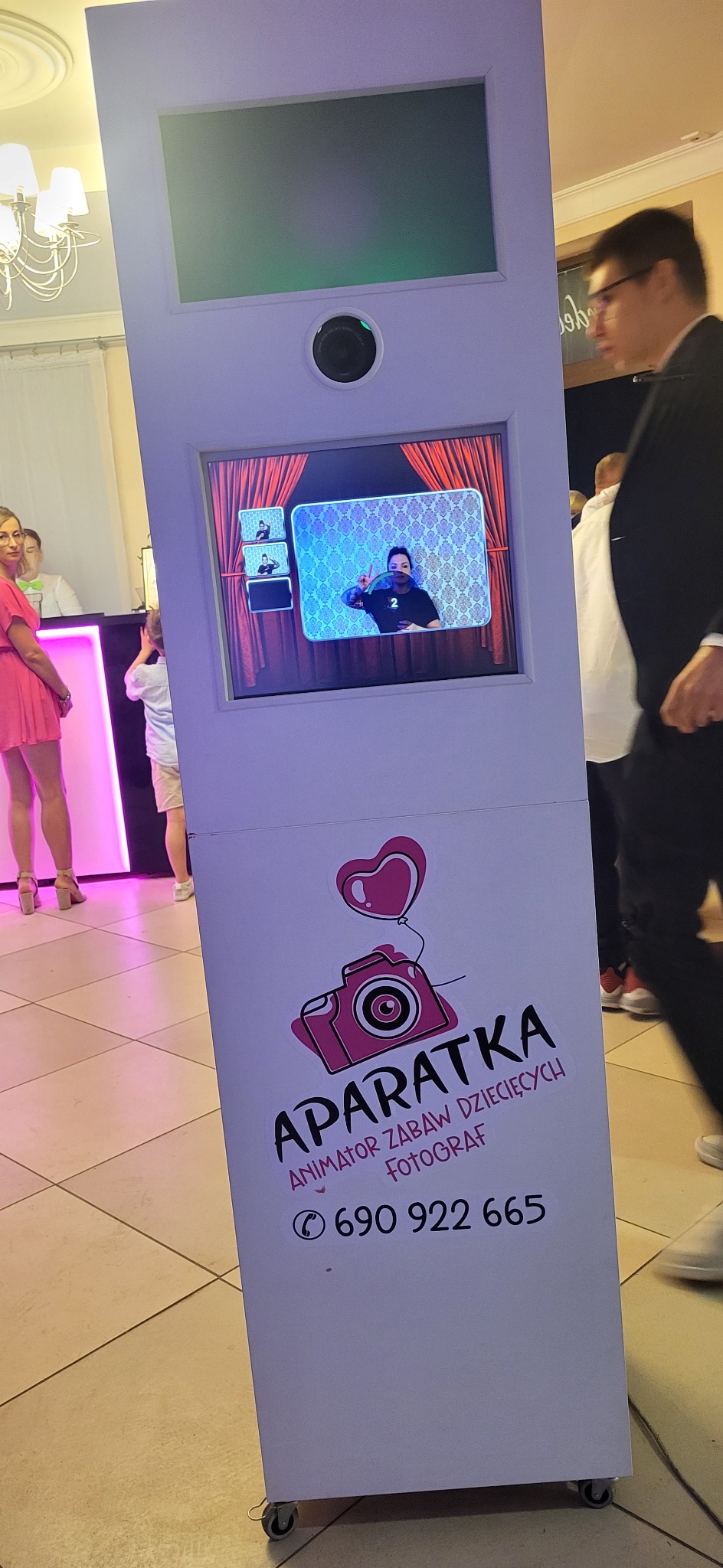 Biała fotobudka z ekranem pokazującym sekwencję zdjęć z czerwonymi zasłonami w tle, logo firmy z aparatem i numerem telefonu widoczne na obudowie, w tle goście na imprezie.