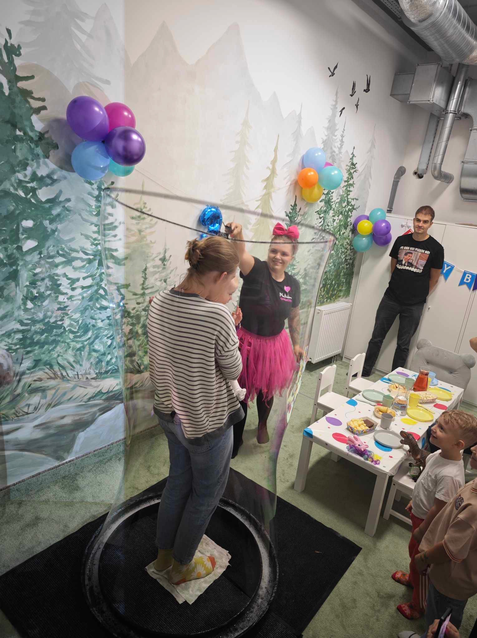 Dziewczynka wewnątrz dużej bańki mydlanej trzymanej przez uśmiechniętą kobietę w różowej spódniczce tutu, w tle dzieci, stół z przekąskami i dekoracje urodzinowe w pokoju z malowidłami leśnymi...