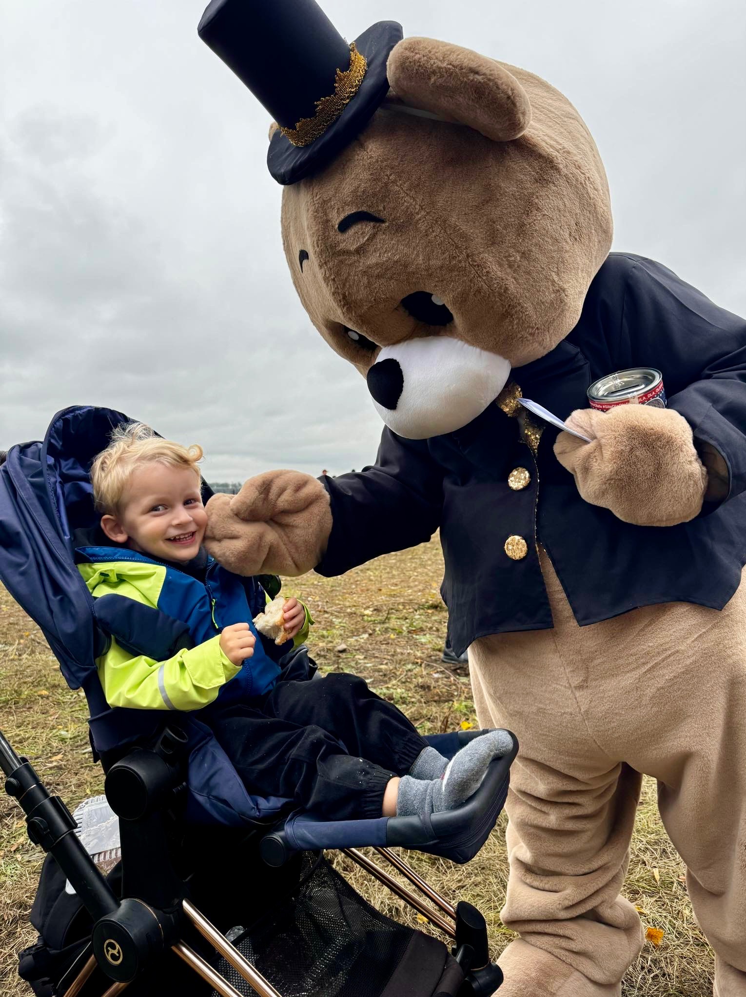 Uśmiechnięty chłopiec w wózku, trzymający kawałek chleba, w interakcji z animatorem przebranym za niedźwiedzia w czarnym fraku i cylindrze, oferującym mu smakołyk z puszki.