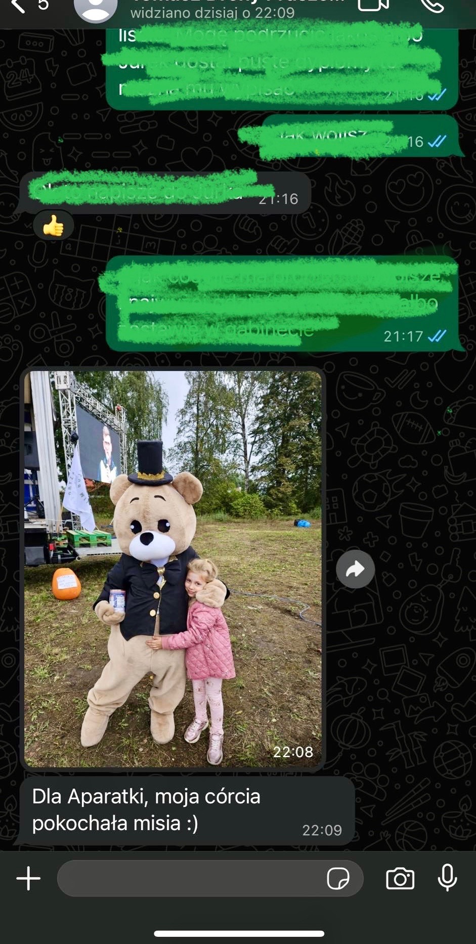 Dziewczynka w różowej kurtce przytula się do animatora przebranego za misia w czarnym fraku i cylindrze na tle zielonego trawnika i sceny.