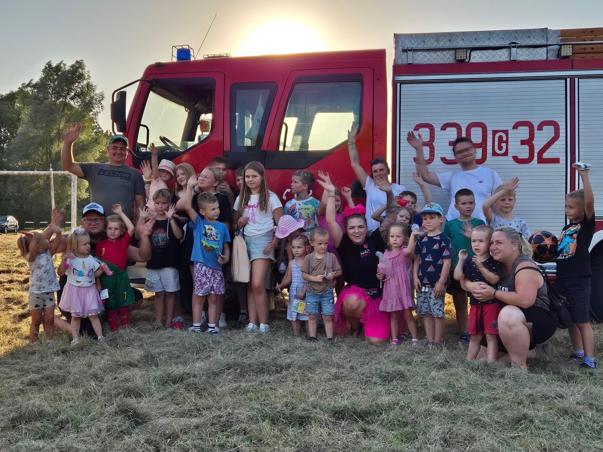 Grupa dzieci i dorosłych macha rękami przed czerwonym wozem strażackim w słoneczny dzień. Kobieta w różowej spódniczce klęczy w środku grupy.